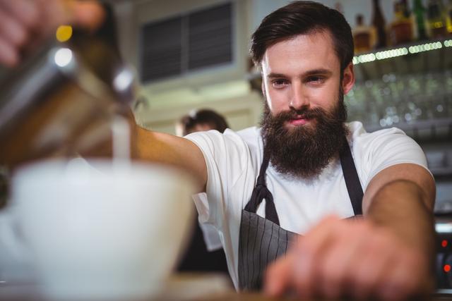 Barista with beard and apron smiling while preparing coffee at cafe counter. Ideal for use in articles or advertisements related to coffee shops, barista training, hospitality industry, and customer service. Perfect for promoting cafes, coffee brands, or barista courses.