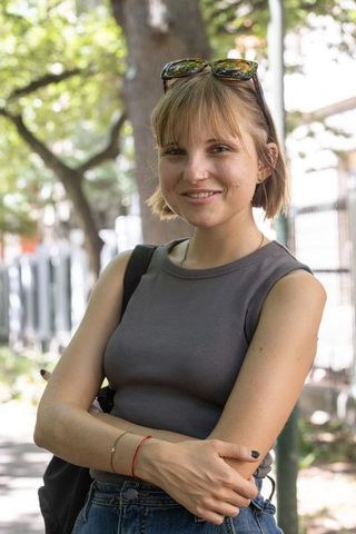 Smiling young woman standing on shaded city sidewalk wearing gray tank top and high waisted denim