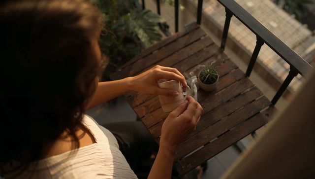 Woman stirring hot drink on sunlit balcony with small succulent and wooden table