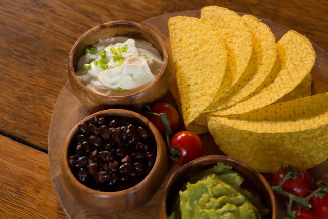 This image features a wooden platter with nachos, guacamole, sour cream, black beans, and cherry tomatoes on a wooden table. Ideal for use in food blogs, restaurant menus, culinary websites, and social media posts promoting Mexican cuisine and festive gatherings.
