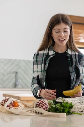 Young woman unpacking groceries at kitchen island holding banana and wearing plaid shirt