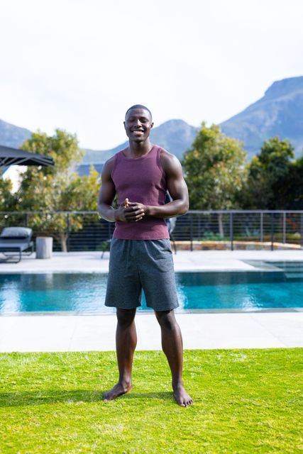 Fit African American man standing barefoot on grass, smiling confidently by a pool in a sunny garden. Ideal for promoting healthy lifestyles, fitness programs, summer activities, outdoor relaxation, and wellness retreats.