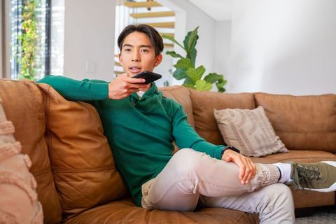 Young asian man relaxing on leather sofa holding remote watching tv in modern living room