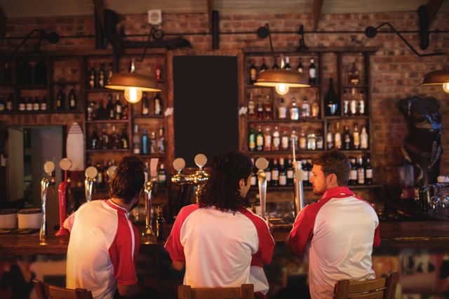 Group of male friends in sports jerseys sitting at a pub counter, enjoying drinks and socializing. Ideal for themes related to friendship, leisure activities, sports fans, nightlife, and social gatherings.
