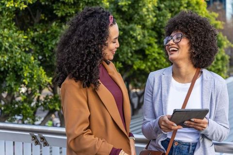 Two women laughing and talking on urban bridge using tablet for casual work talk