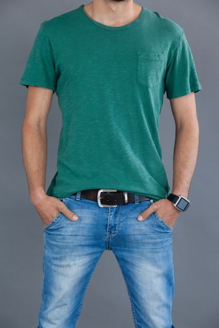 Man wearing a green t-shirt and blue jeans, posing with hands in pockets against a grey background. Ideal for fashion, casual wear promotions, lifestyle blogs, and clothing advertisements.
