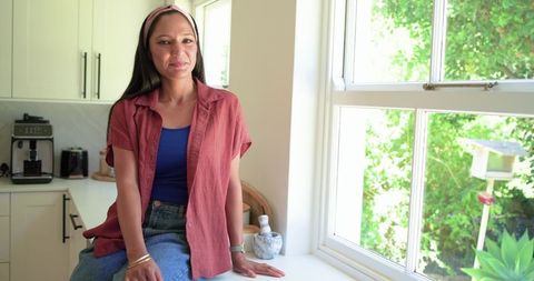 African woman sitting on white kitchen counter by sunny window wearing pink headband
