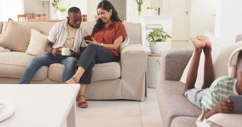 Couple sharing smartphone on sofa while child using tablet in bright modern living room