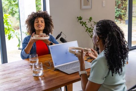 Two african american women sharing headphones while testing laptop audio in home workspace
