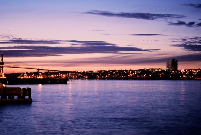 This mesmerizing scene captures a waterfront during dusk, with the twinkling lights of the city skyline reflecting off the water. The image features a bridge spanning across the horizon, against a backdrop of a vibrant, colorful twilight sky. Ideal for use in travel brochures, cityscape artwork, websites promoting urban destinations, or for articles on serene waterfront views and urban beauty.