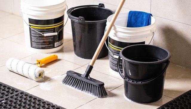 Janitorial supplies on beige tile floor with push broom, buckets, roller and blue cloth