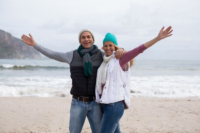 Mature couple standing on the beach with arms outstretched, dressed in winter clothing. They are smiling and appear happy, enjoying their time together by the ocean. This image is perfect for use in travel brochures, lifestyle blogs, advertisements for winter beach vacations, or articles about maintaining a happy relationship in later years.