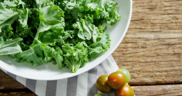 Fresh kale leaves are arranged on a white plate, with a rustic wooden background and a striped cloth adding to the organic feel, with copy space. Cherry tomatoes provide a pop of color alongside the vibrant green, suggesting a healthy and nutritious meal preparation.