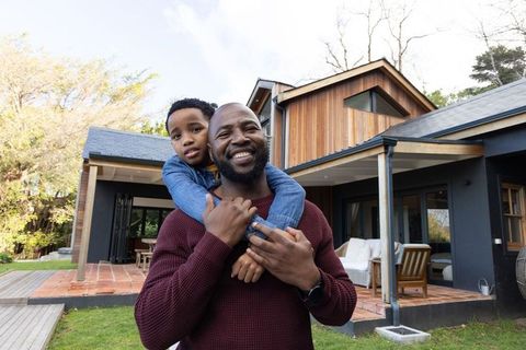 African american father carrying son piggyback smiling outside modern suburban home