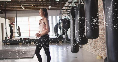 Woman standing in boxing gym clasping hands by punching bags backlit industrial studio