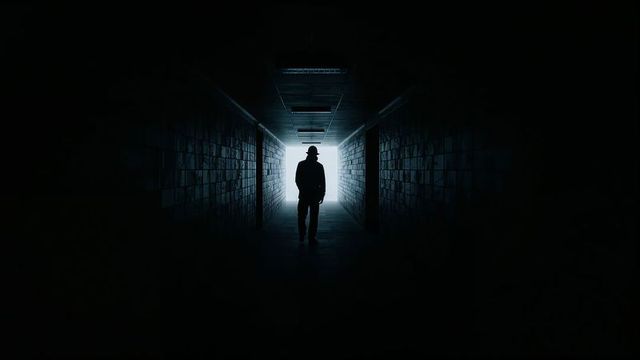 Silhouette of man with hat walking through dark corridor toward bright exit