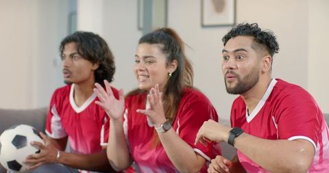 Diverse friends cheering and reacting while watching soccer on couch wearing red jerseys at home