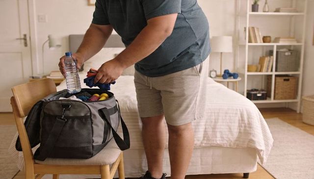 Packing gym duffel bag at home: man organizing water bottle, towels, dumbbells for workout