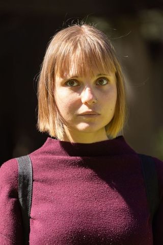 Young woman wearing maroon sweater and nose ring, standing in dappled sunlight, calm expression