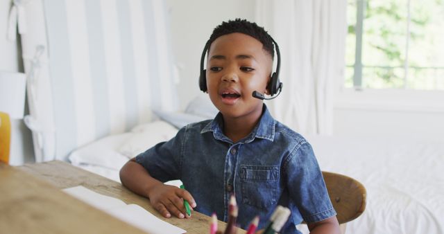 Young African American boy wearing a headset sits at a desk studying online in a bright, well-lit room. Perfect for use in articles or advertisements related to remote education, e-learning, childhood learning activities, technology in education, and family-friendly content.