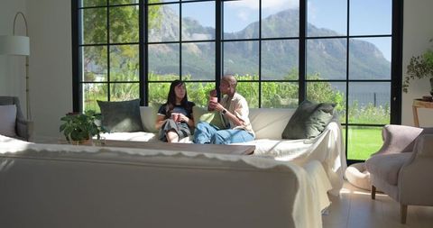 Two women sitting on sofa by floor-to-ceiling windows with mountain view holding mugs