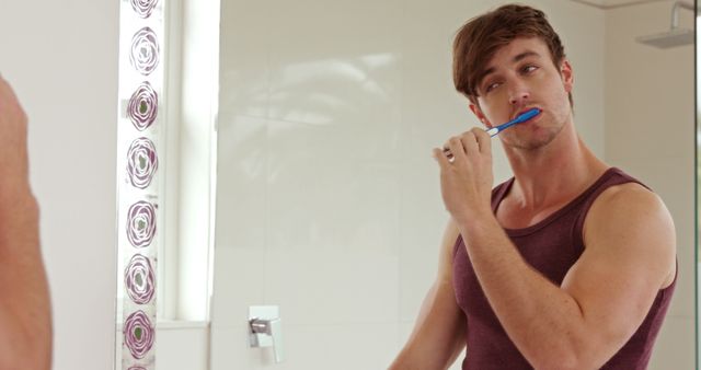 Young man brushing teeth in front of bathroom mirror. Casual outfit, reflecting self-care and hygiene. Ideal for topics on daily routines, personal care, or morning rituals in health and lifestyle contexts.