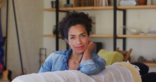 A woman in a denim shirt is sitting on a couch at home, looking relaxed and comfortable. She has one hand resting on her head and is smiling softly. In the background, there are shelves with books and decor items. This image shows relaxation, domestic life and calm. Perfect for use in articles about home living, personal reflections, positivity, lifestyle blogs, and mental well-being.