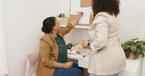 Women collaborating at desk pointing at corkboard during creative planning session