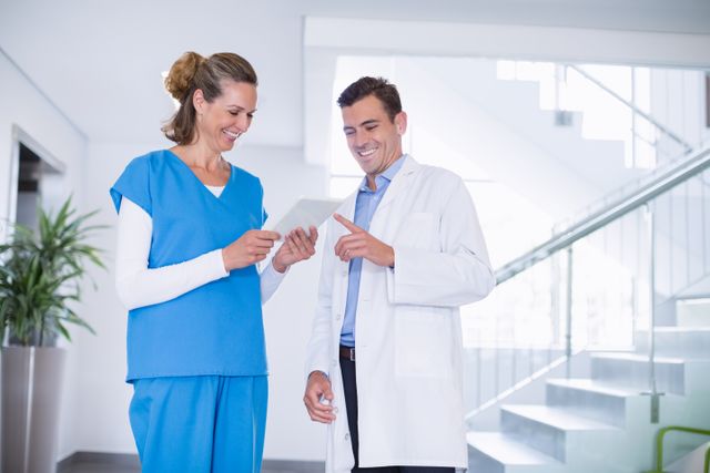 Nurse and doctor standing in a hospital corridor, engaged in a discussion over a digital tablet. Both are smiling, indicating a positive and collaborative work environment. This image can be used for healthcare-related content, medical teamwork, modern healthcare technology, and professional collaboration in medical settings.