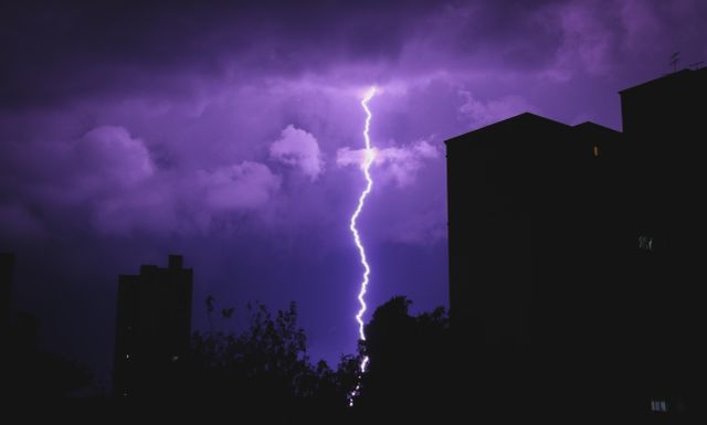 This dramatic scene of a bright lightning bolt piercing a purple night sky over a cityscape silhouette captures natural power and beauty. Use it for weather-related features, dramatic visuals, nature articles, or presentations on urban environments.