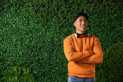 Asian man standing against green foliage wall in orange sweater and headphones