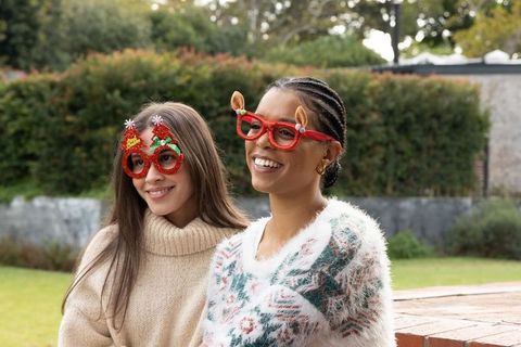 Diverse women wearing festive novelty glasses and cozy sweaters smiling in backyard