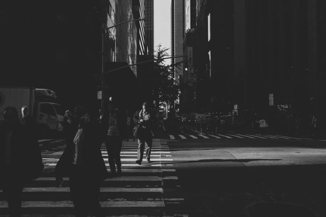 This moody urban scene features pedestrians emerging from shadows as they cross a busy city intersection in the early morning light. Ideal for concepts of daily commute, urban living, city life, and metropolitan atmosphere.