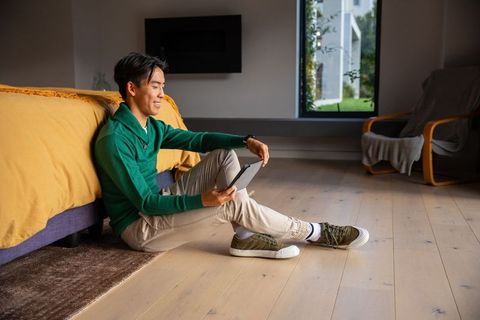 Asian man sitting on bedroom floor using tablet smiling in cozy sunlit modern bedroom