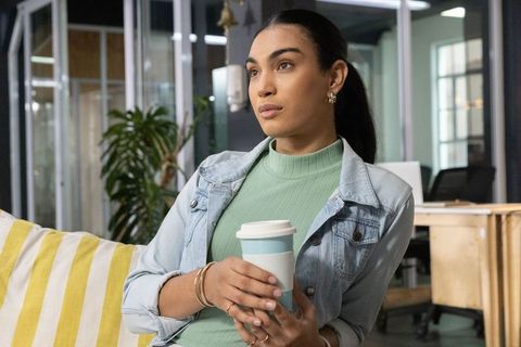 Young woman in denim jacket holding reusable coffee cup in coworking lounge, contemplative