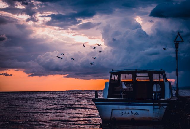 This beautiful and serene photo captures a boat anchored at sunset with birds flying over the ocean. Ideal for travel brochures, nature and landscape blogs, or any content aimed at promoting peaceful and tranquil environments. The dramatic clouds and vibrant colors of dusk create a captivating visual impact.