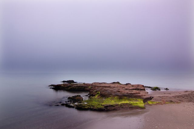 The image showcases a tranquil rocky beach with calm water and a misty purple sky, creating a serene and peaceful atmosphere. This beautiful coastal scenery is perfect for use in travel brochures, nature blogs, and relaxation-themed projects. The smooth transition between the sea and sky enhances its soothing qualities, making it ideal for backgrounds, wallpapers, and posters promoting tranquility and meditation.