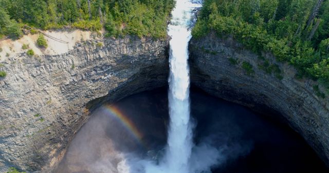 Aerial view captures a majestic waterfall cascading into a circular basin, creating a misty rainbow in the natural amphitheater. The geological formations surrounding the waterfall highlight the raw power and beauty of nature.