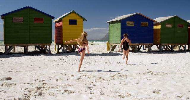Happy caucasian girls in swimsuits running near wooden houses on sunny beach. Childhood, friendship, free time, summer, travel, vacations and lifestyle, unaltered.
