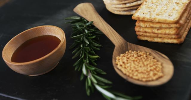 A wooden bowl of honey is accompanied by crackers, fresh rosemary, and a spoonful of sesame seeds on a dark surface. These ingredients suggest a setting for a culinary preparation or a gourmet food presentation.