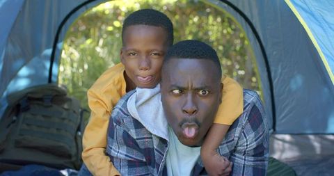 Father and son making funny faces while camping in tent, playful outdoor family bonding