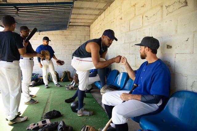 Multi ethnic baseball team preparing for a game in a locker room. Two players are fist bumping, showcasing camaraderie and teamwork. Ideal for content related to sports, teamwork, athletic preparation, and team spirit.