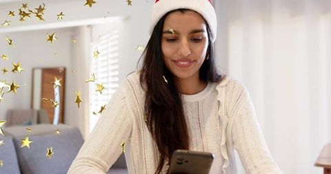 Indian woman wearing santa hat scrolling phone in cozy home with golden stars overlay