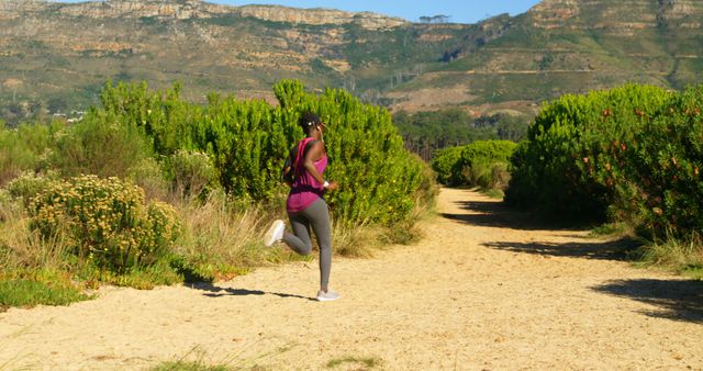 This image shows a woman running on a trail surrounded by lush greenery and mountains in the background on a sunny day. This can be used for promoting outdoor fitness, healthy lifestyle activities, exercise rehabilitation, trekking destinations, motivational health posters, and eco-tourism.