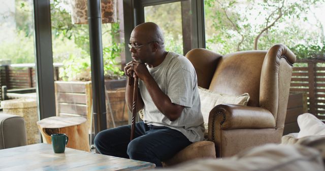 Thoughtful african american senior man sitting in living room leaning on walking stick. retirement lifestyle, spending time alone at home.