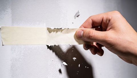 Hand pulling masking tape from painted wall revealing plaster flakes and texture
