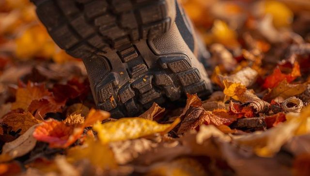 Hiking boot tread pressing through autumn leaves on forest trail, close-up rugged texture