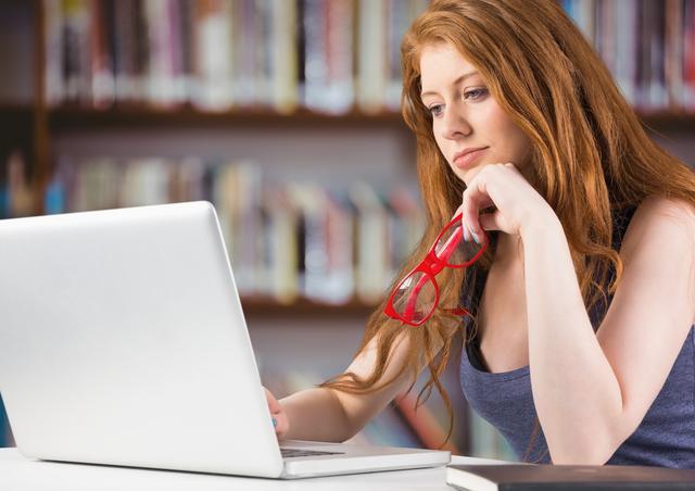 Young woman with red hair holding glasses while using laptop in library. Ideal for educational content, online learning platforms, academic research, and technology in education themes.