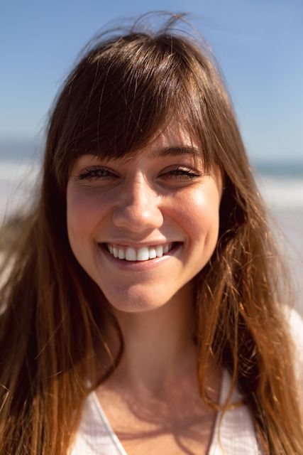 Young woman smiling at camera with sunlight on her face, enjoying a day at the beach. Ideal for concepts of happiness, carefree living, summer vacations, and outdoor enjoyment.