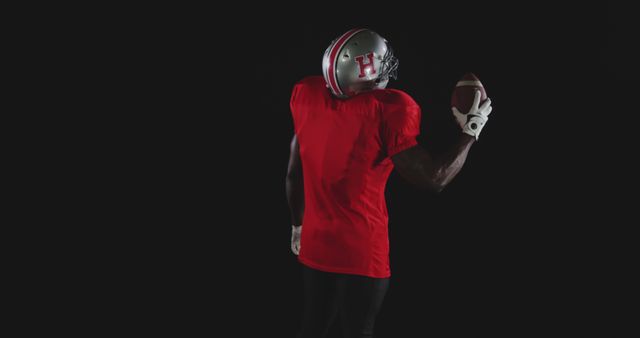 Image showcases an American football player wearing red jersey, gripping a football in one hand with black background. Ideal for sports-related advertisements, training materials, team promotions, or competitive sports event posters.